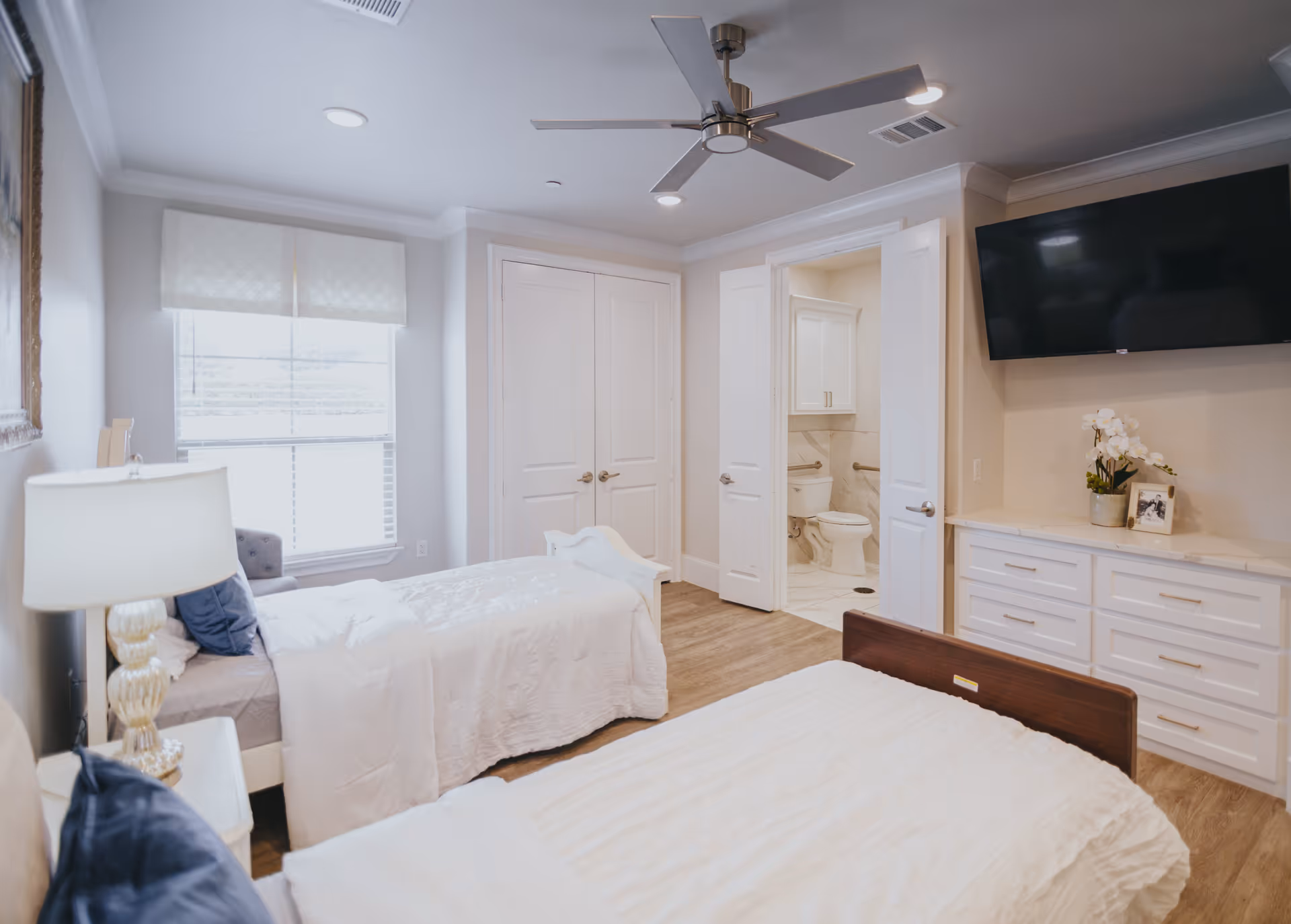 A bright and clean bedroom with two single beds, white bedding, and blue pillows. There is a bedside table with a lamp, a large window with a white valance, a ceiling fan, a wall-mounted TV above a white dresser, and an open door leading to a bathroom with a toilet and grab bars.