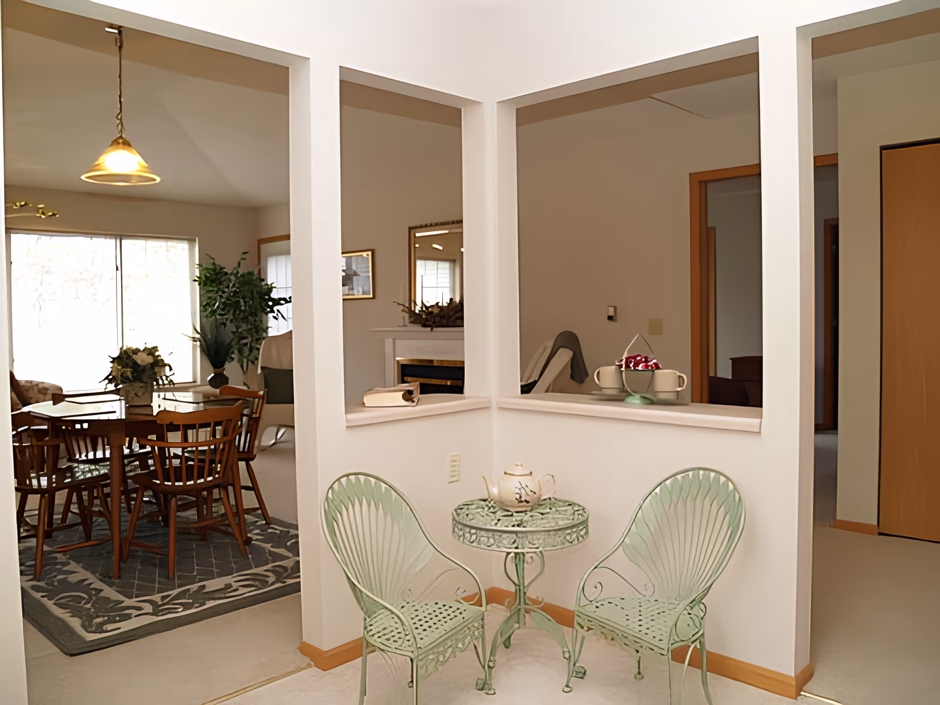 A cozy interior space featuring a small green metal table with two matching chairs. On the table is a teapot. The room has three large windows or openings in the wall, showing a dining area with a wooden table and chairs, a plant, and a fireplace in the background. The walls are light-colored and the floor is carpeted.