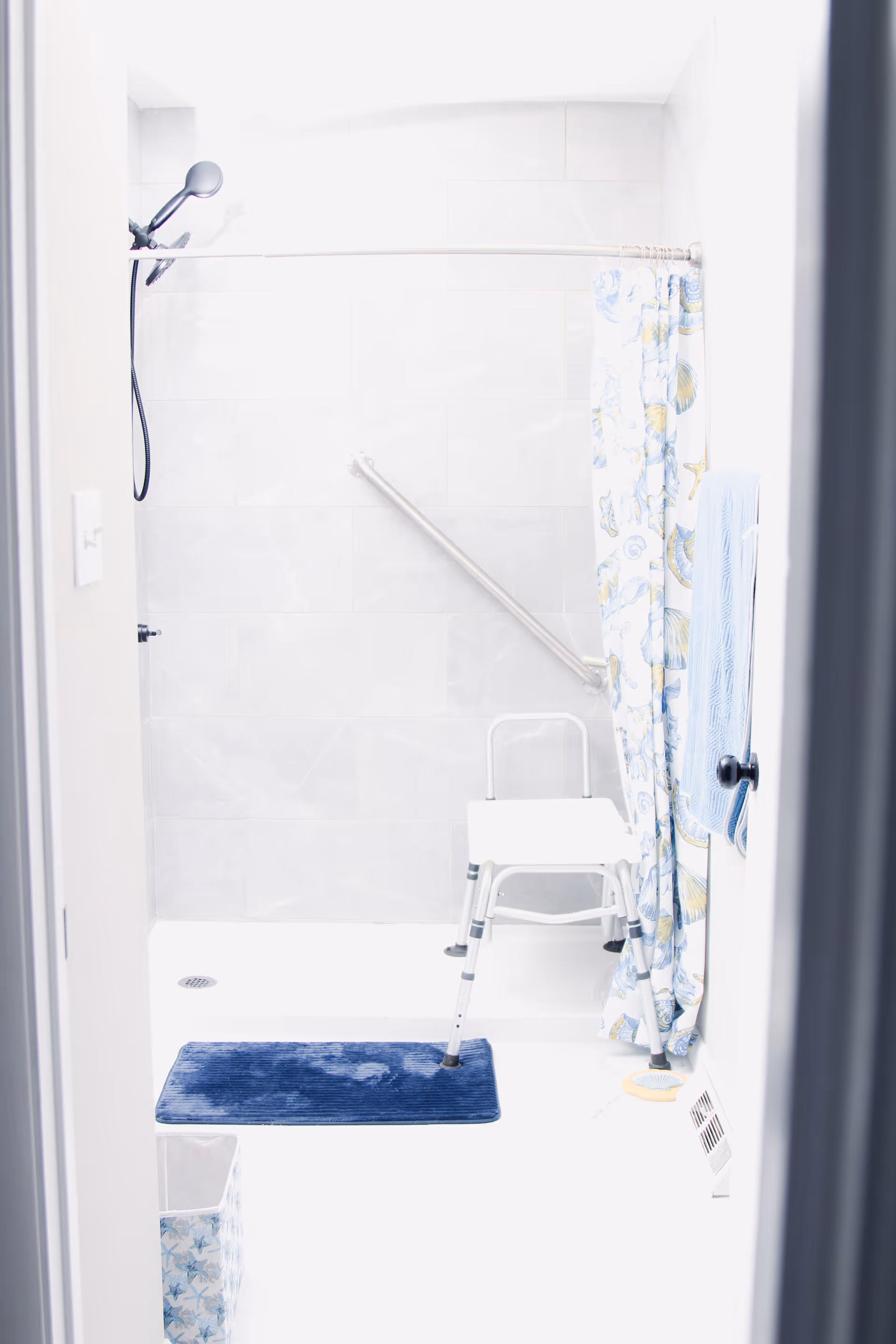 A bright bathroom with a walk-in shower featuring a handheld showerhead, a grab bar on the tiled wall, a white shower chair, a blue bath mat on the floor, and a shower curtain with a seashell pattern. A blue towel hangs on a rack beside the shower.