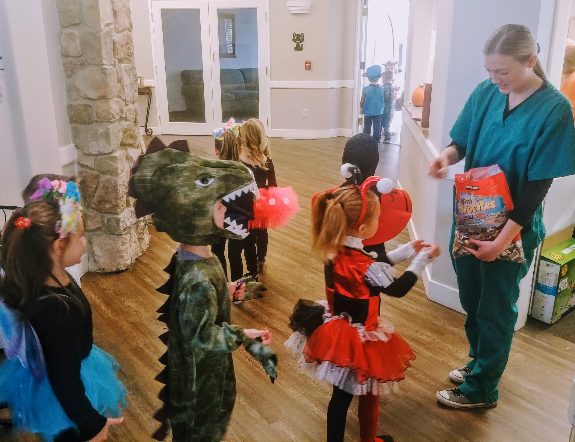 A group of young children dressed in various Halloween costumes, including a dinosaur, ladybug, and fairy, standing in a hallway of a senior living facility. A staff member in green scrubs is handing out candy from a large bag to the children. The hallway has wooden floors, stone pillars, and white walls with doors and windows in the background.