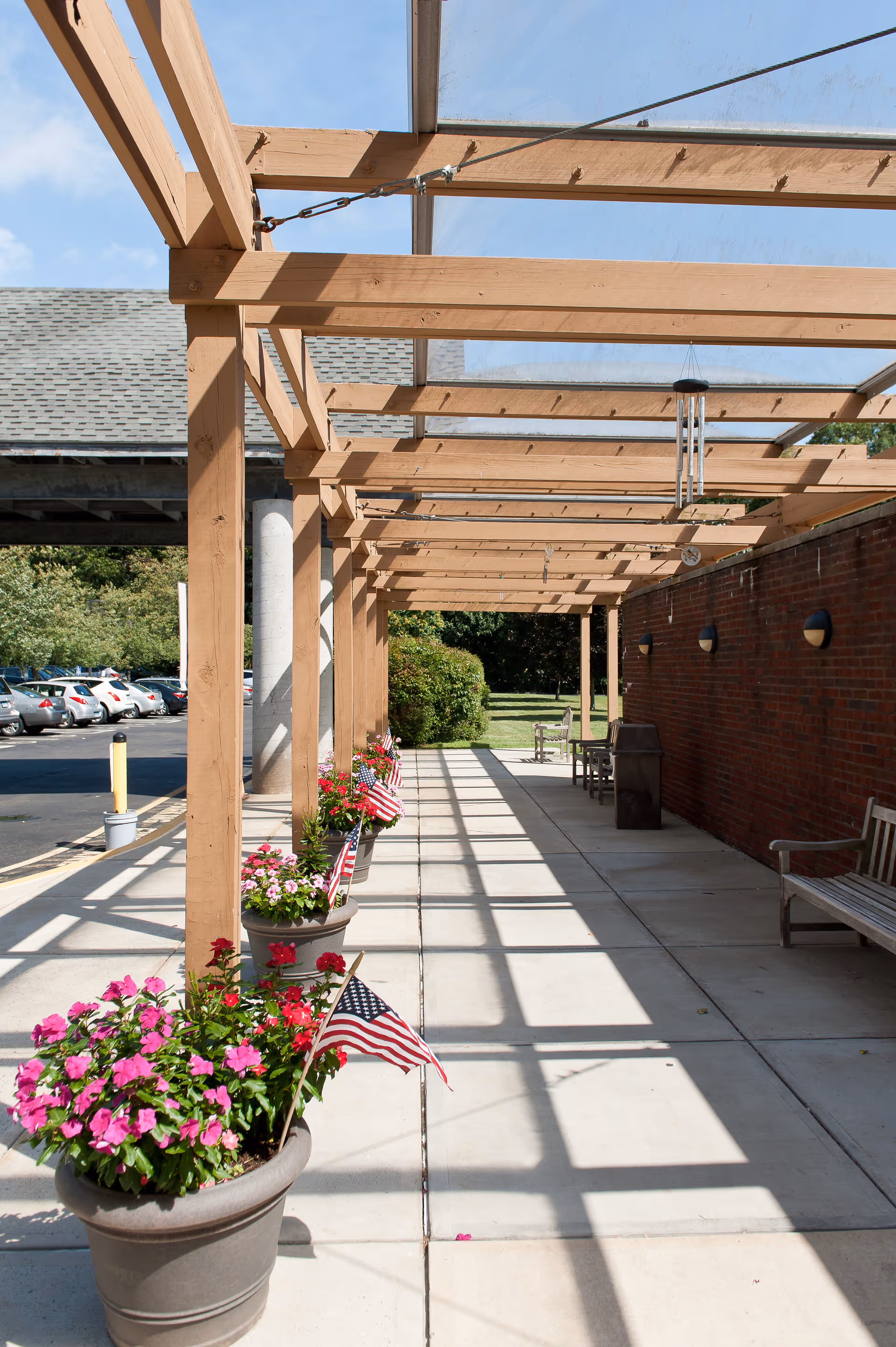 Shaded outdoor walkway with a wooden pergola, potted flowers and small American flags along a brick wall with benches.