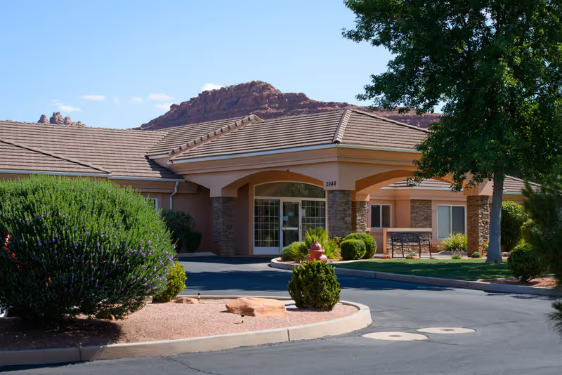 Exterior view of a single-story assisted living facility with a covered entrance, surrounded by landscaped bushes and trees, with red rock formations visible in the background under a clear blue sky.
