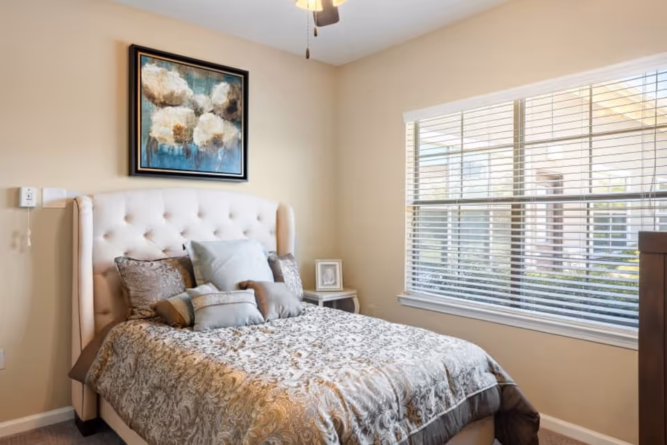 A cozy bedroom with a tufted beige headboard bed adorned with multiple decorative pillows and a patterned comforter. A framed floral painting hangs above the bed on a beige wall. To the right, there is a large window with white blinds letting in natural light. A small side table with a framed photo is next to the bed.
