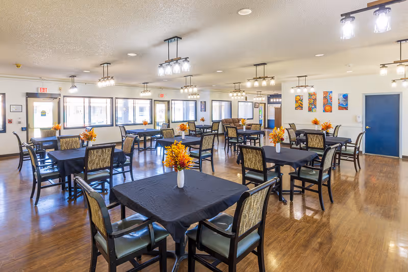 A spacious dining room with multiple square tables covered with black tablecloths, each adorned with a small vase of orange flowers. The room has wooden flooring, several windows letting in natural light, modern ceiling lights, and colorful artwork on one wall.