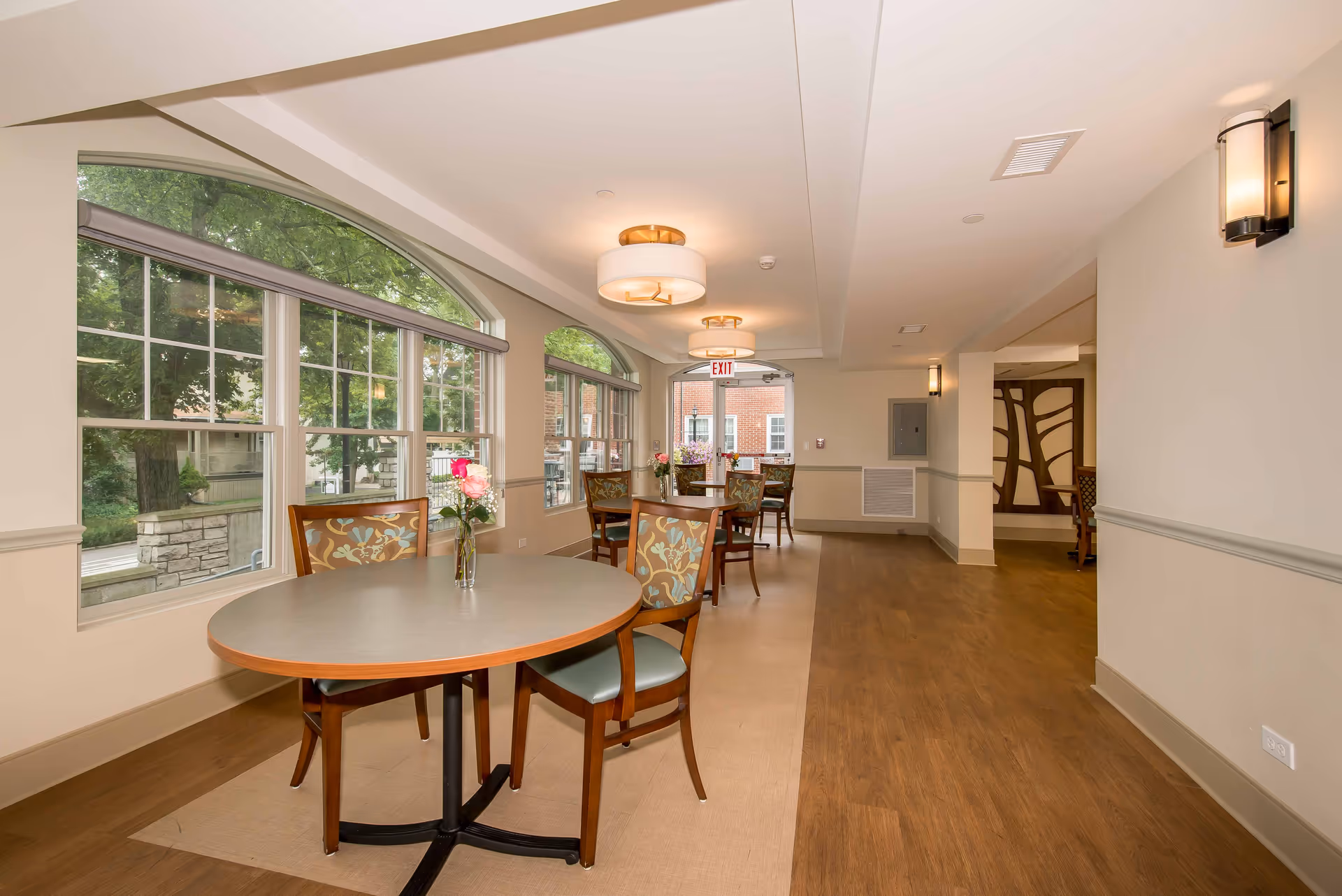 Interior view of a senior living facility dining area with round tables and chairs. The room has large arched windows letting in natural light, light-colored walls, and wood flooring. Each table has a small vase with flowers. Ceiling lights and wall sconces provide additional lighting.