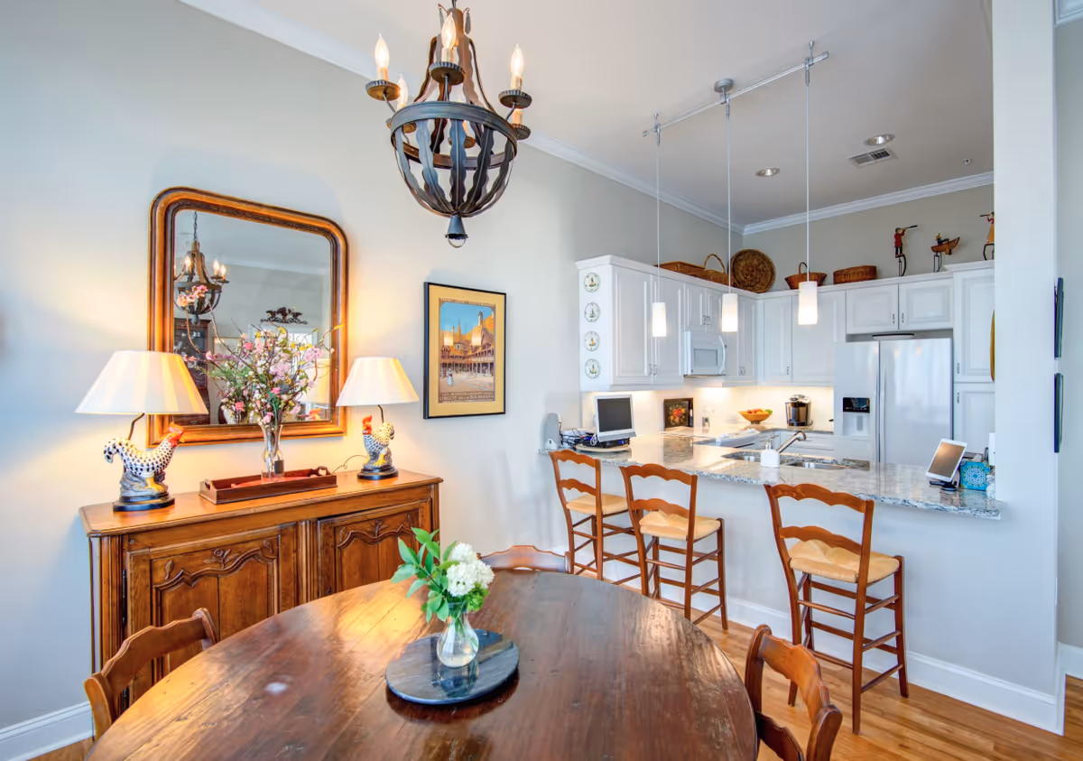 A bright and cozy dining area with a wooden round table and chairs, a wooden sideboard with two lamps and a large mirror above it. Adjacent to the dining area is a modern kitchen with white cabinets, granite countertops, a refrigerator, microwave, and three wooden bar stools at the counter. Pendant lights hang above the kitchen counter, and decorative items are placed on top of the cabinets.