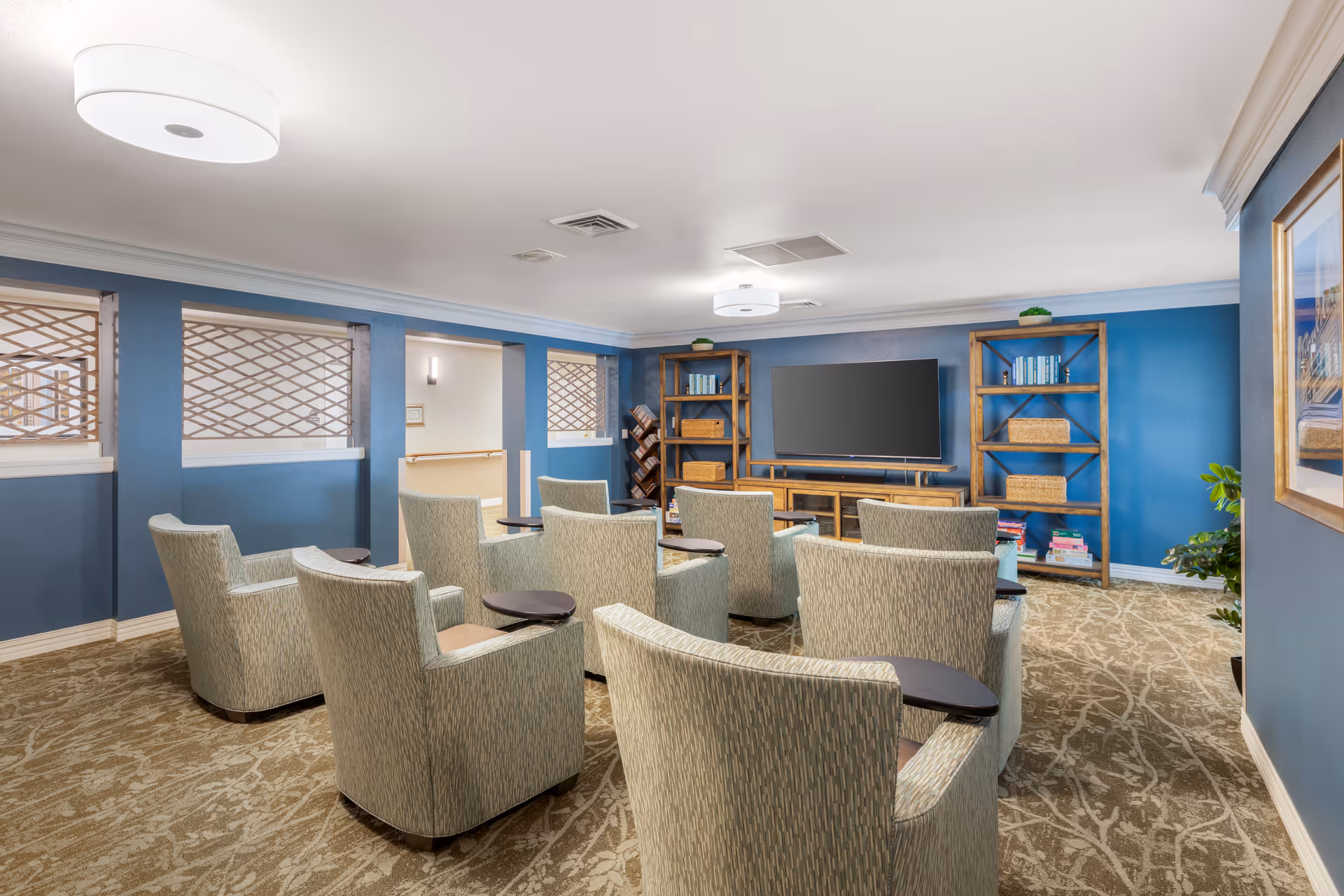 Community lounge with several upholstered armchairs arranged in rows facing a large flat-screen TV and wooden shelving against blue-painted walls.