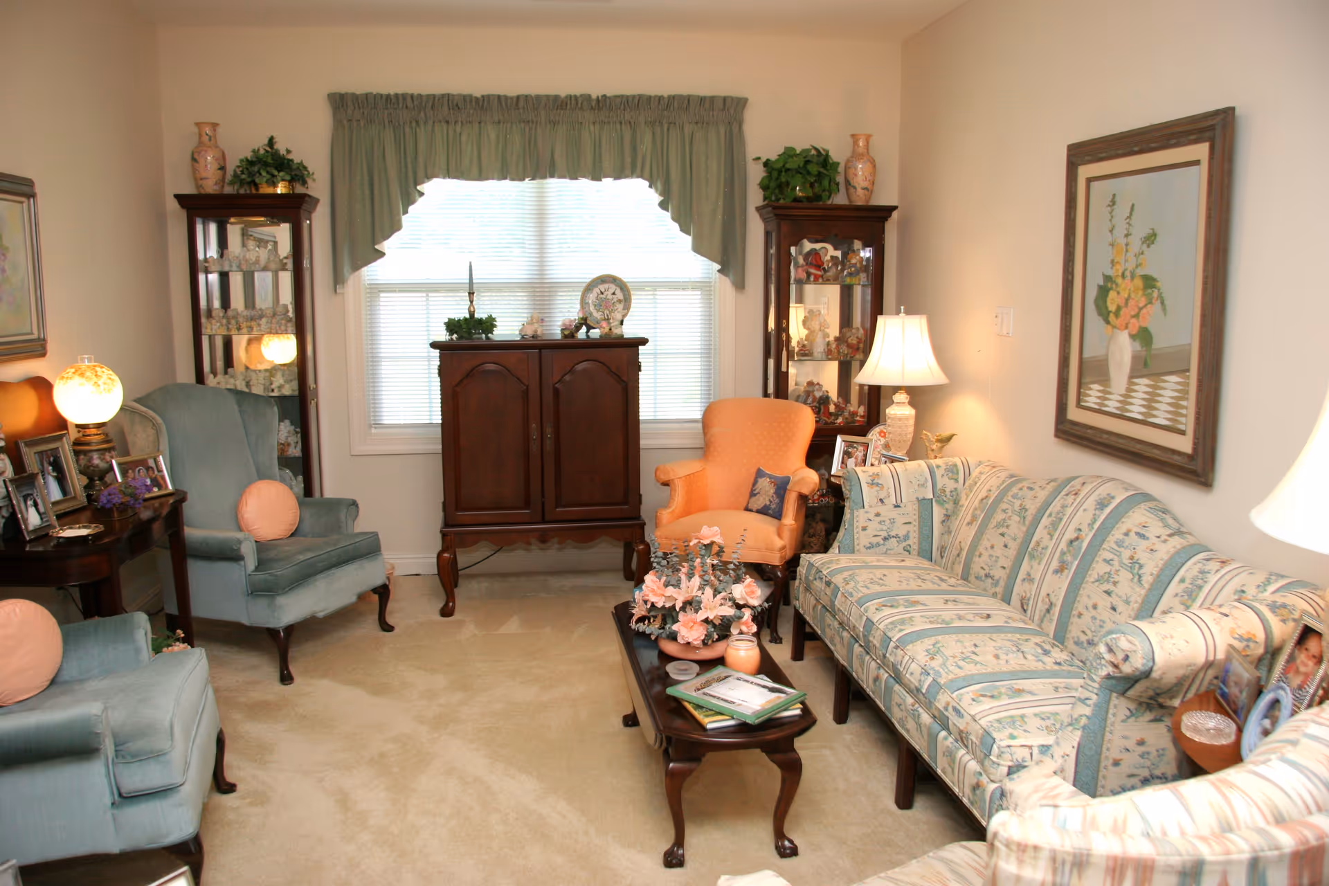 A cozy living room with a floral patterned sofa, two blue armchairs with round pink cushions, and an orange armchair. There is a wooden coffee table with a flower arrangement and some books. Two glass display cabinets with decorative items flank a window with green valance curtains. The room is softly lit by table lamps and decorated with framed pictures and a floral painting on the wall.
