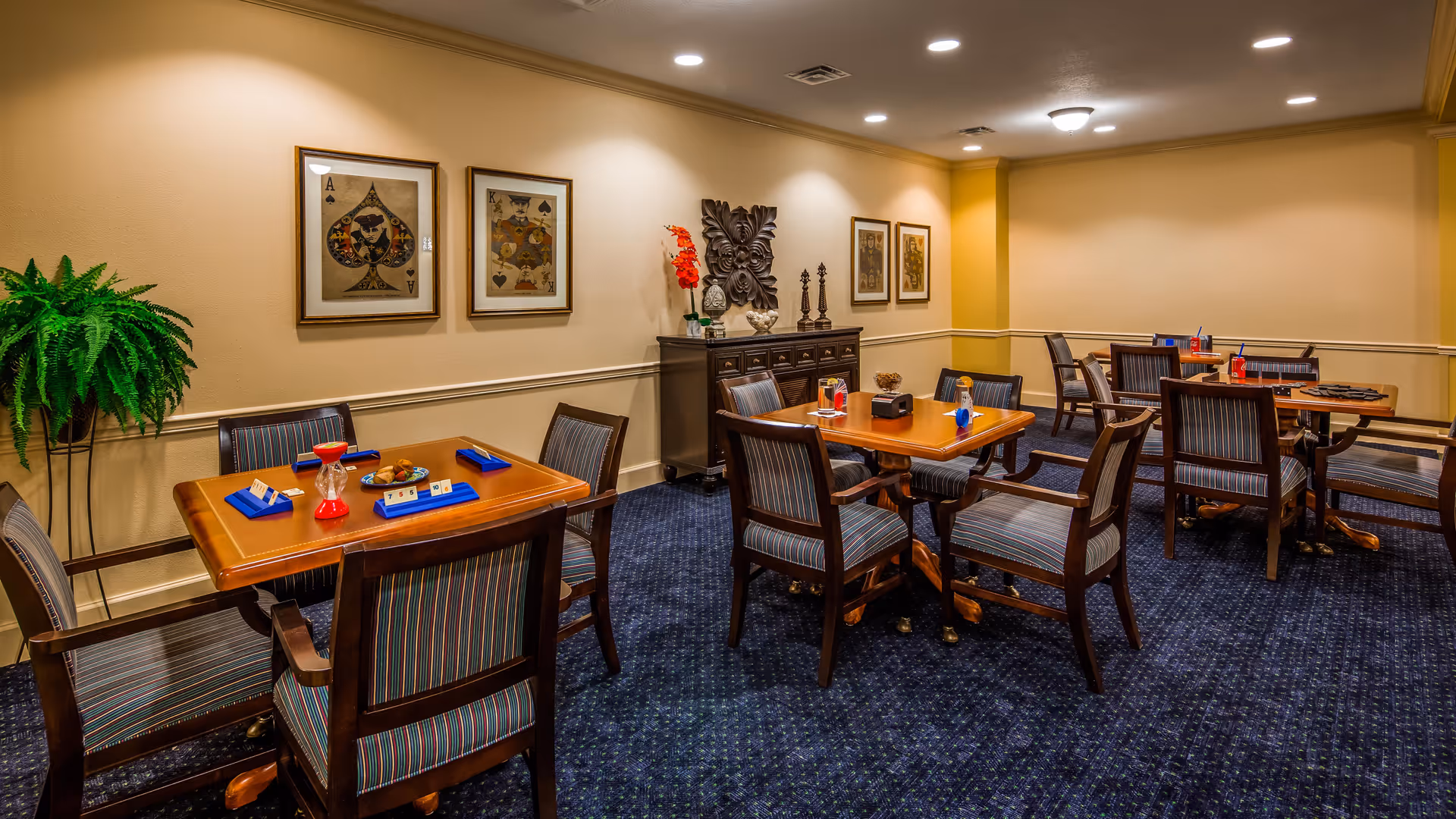 A well-lit room with several wooden tables and striped upholstered chairs arranged for group activities or dining. The tables have game timers, scorecards, and small decorative items. The walls are beige with framed artwork and a decorative cabinet with sculptures and flowers. A green potted plant is in the corner.