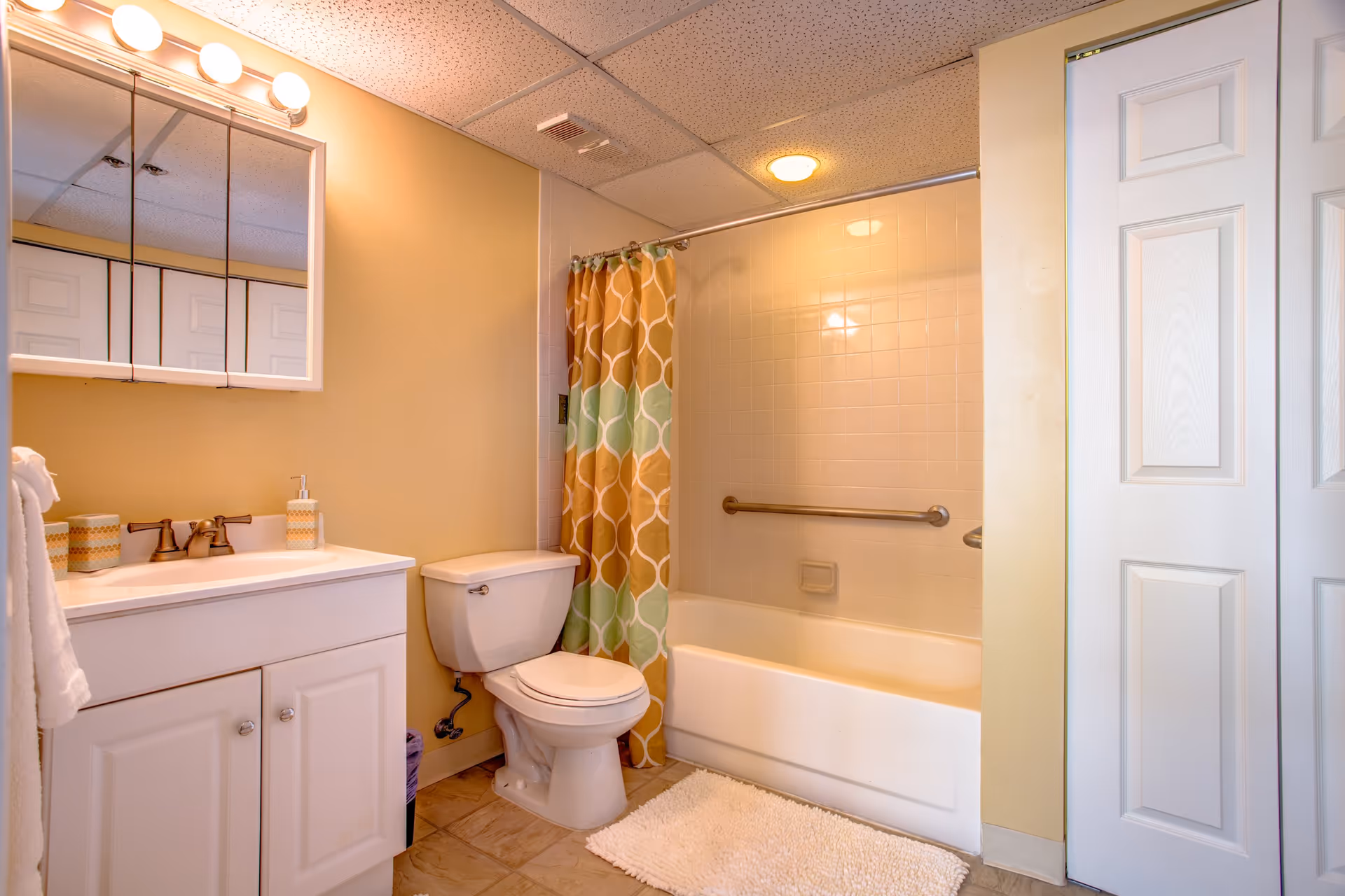 A bathroom with a white vanity and sink, a toilet, and a bathtub with a colorful shower curtain in shades of orange, green, and white. There is a white bath mat on the tiled floor, a towel hanging on the left side, and a mirrored medicine cabinet above the sink. The walls are painted light yellow and the ceiling has white tiles with a light fixture above the mirror.