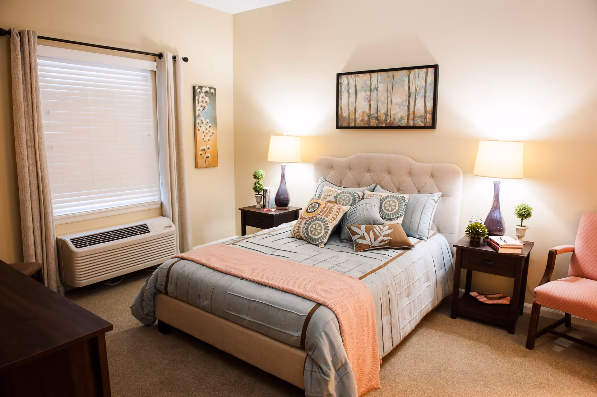 A cozy bedroom with a beige upholstered bed adorned with multiple decorative pillows and a light blue and beige bedspread with a peach throw blanket. There are two dark wooden nightstands on either side of the bed, each with a lamp and small decorative plants. A pink upholstered chair is positioned to the right, and a window with beige curtains and white blinds is on the left wall. The walls are painted a soft beige color and decorated with two pieces of artwork.