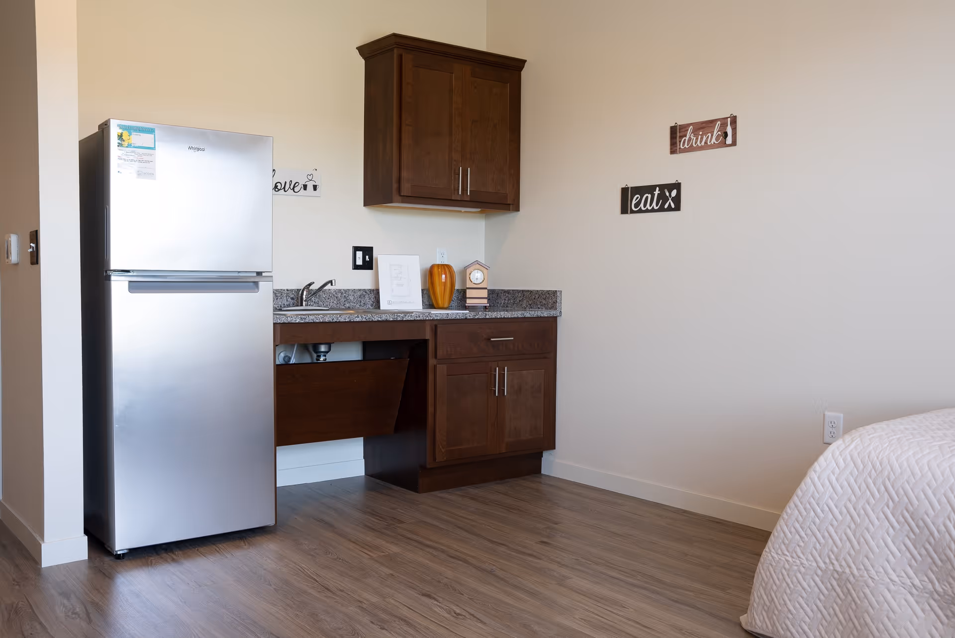 Small kitchenette area with a stainless refrigerator, wooden cabinets, a sink and a partial bed visible at right.