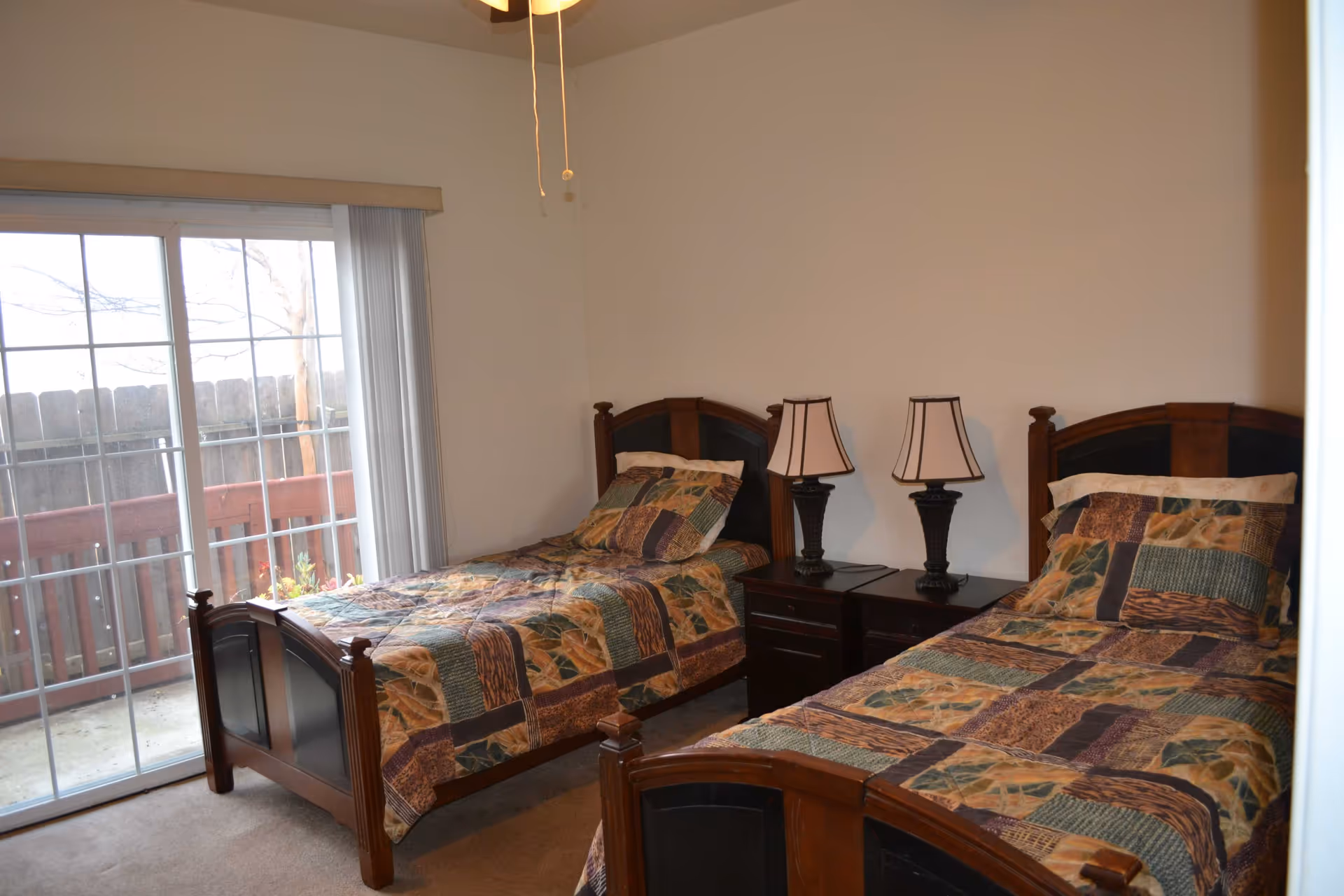 A bedroom with two twin beds featuring wooden headboards and patchwork quilts. Between the beds is a dark wooden nightstand with two matching table lamps. A sliding glass door with vertical blinds leads to an outdoor area with a wooden fence and deck.