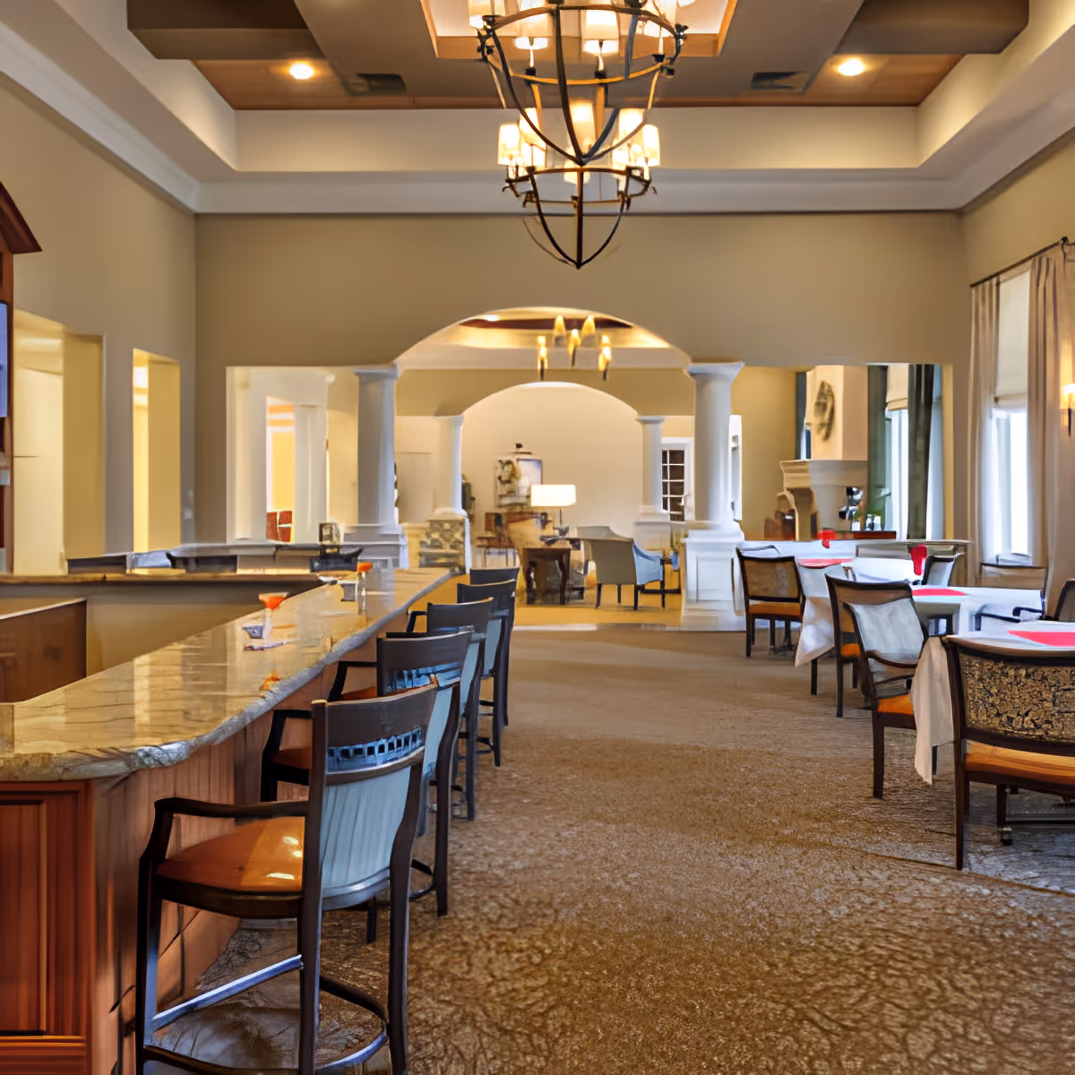 Interior view of a spacious dining area with a long marble countertop bar on the left side, lined with high chairs. On the right side, there are multiple dining tables with chairs, some tables set with red placemats. The room features high ceilings with decorative lighting fixtures and large windows with curtains allowing natural light to enter. In the background, there is an archway supported by columns leading to another seating area with armchairs and a lamp.