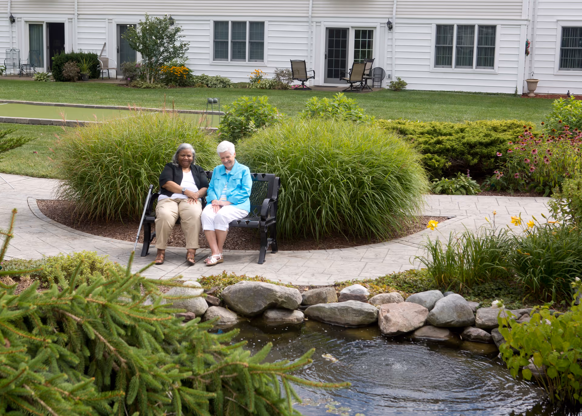 Two elderly women sitting on a bench in a garden area with lush greenery, flowers, and a small pond with rocks in the foreground. Behind them is a white building with windows and a patio area with chairs and a table.