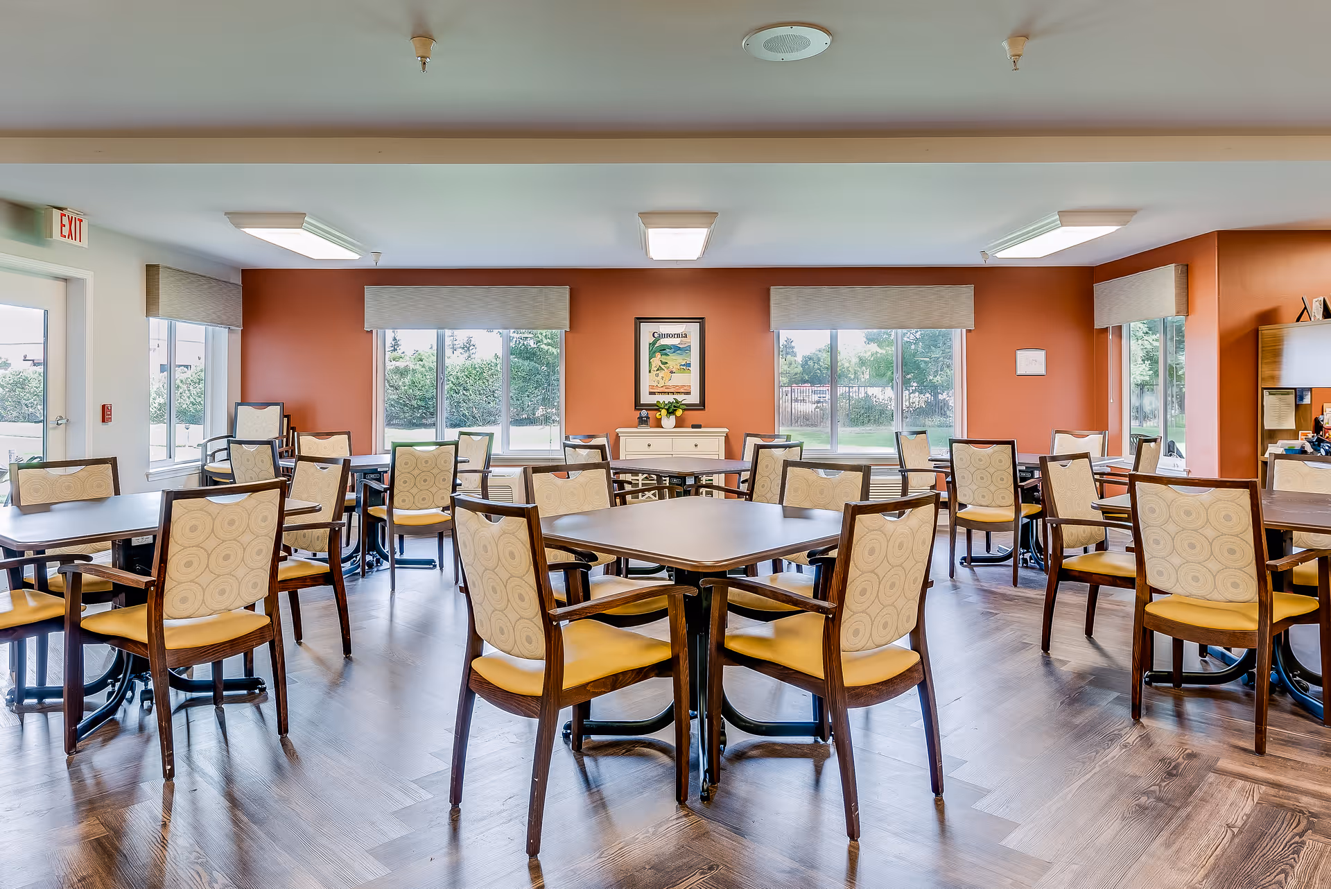 A bright dining room with multiple square tables and wooden chairs with patterned cushions. The room has large windows with beige valances, allowing natural light to fill the space. The walls are painted a warm orange color, and there is a framed picture and a small cabinet against the far wall. The floor is a dark wood laminate.
