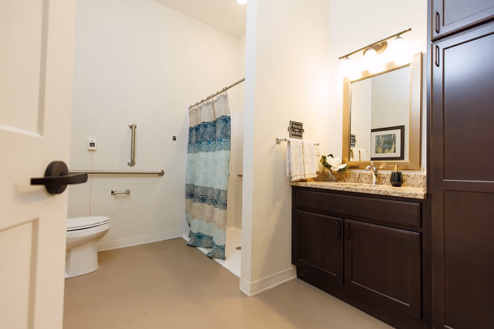 A clean and accessible bathroom with a toilet, grab bars, a shower with a blue and white patterned curtain, and a vanity with a granite countertop, dark wood cabinets, a mirror, and a light fixture above. A small sign above the towel rack reads 'Wash Your Hands'.