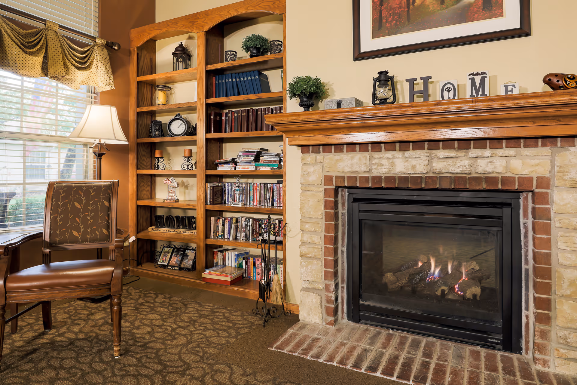 Cozy living room area with a lit fireplace surrounded by a stone and brick mantel. To the left of the fireplace is a wooden bookshelf filled with books, DVDs, and decorative items. A wooden chair with patterned upholstery sits near a window with blinds and a valance. A table lamp is also visible next to the chair, casting warm light in the room.