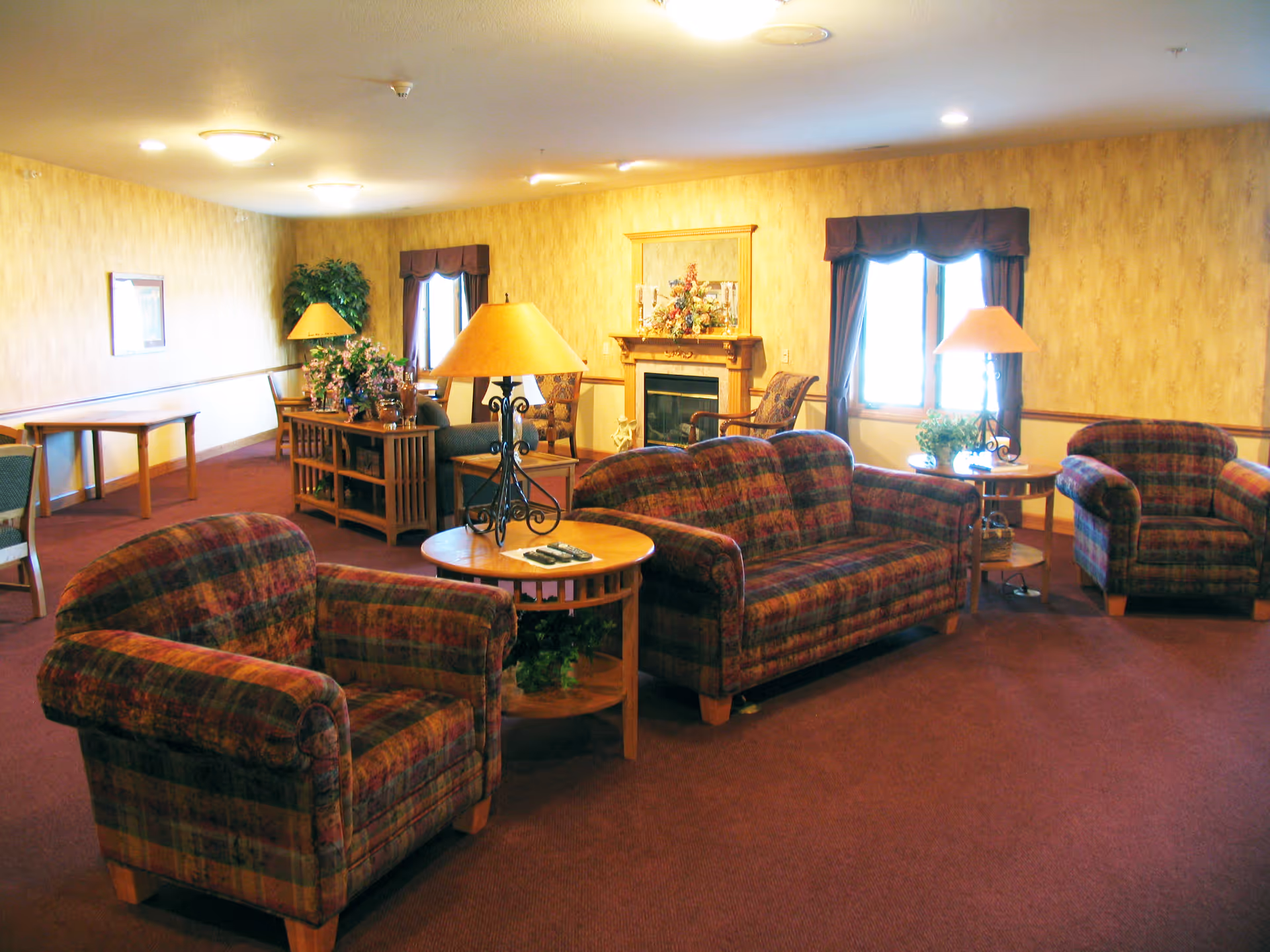 A cozy living room area with plaid upholstered armchairs and a sofa arranged around wooden side tables with lamps. There is a fireplace with a floral arrangement on the mantel, two windows with dark curtains, and a wooden console table with decorative plants. The walls are covered with light wallpaper and the floor is carpeted in a warm tone.