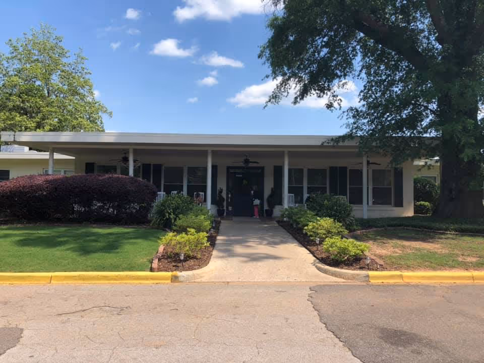 Front exterior view of a single-story building with a covered porch, white rocking chairs, and a walkway leading to the entrance. The building is surrounded by green grass, bushes, and large trees under a partly cloudy blue sky.