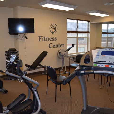Interior view of a fitness center with exercise equipment including a treadmill, stationary bike, weight bench, and rowing machine. There are chairs arranged in the room, a wall-mounted TV, and large windows with blinds letting in natural light. The wall has the text 'Fitness Center' with a logo above it.