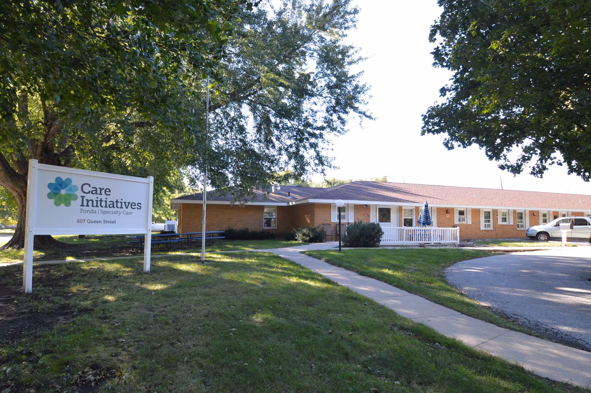Exterior view of a single-story brick building with a white porch and a parked vehicle. There is a large sign in the foreground that reads 'Care Initiatives Fonda | Specialty Care 607 Queen Street'. The area is surrounded by green grass and large trees.