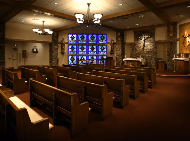 Interior view of a chapel with wooden pews arranged in rows facing an altar. The altar area features religious statues, a crucifix mounted on a stone wall, and stained glass windows with blue and purple designs. The ceiling has wooden beams and hanging light fixtures.