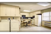 A clean and well-lit laundry and kitchen area featuring a washing machine and dryer on the left, a small dining table with four chairs in the center, and a kitchen with cabinets, microwave, stove, and refrigerator along the back wall. A window with blinds and an air conditioning unit are visible on the right side.