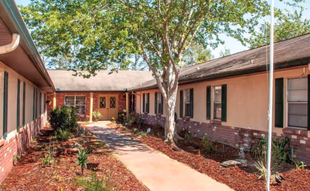 Outdoor courtyard area of a senior living facility with a concrete walkway, landscaped garden beds with mulch and plants, and a large tree in the center. The building has beige walls with brick accents and green window shutters.