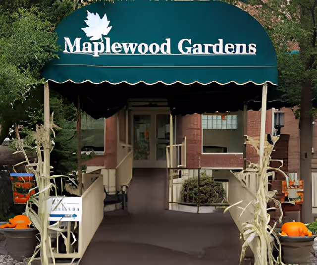 Entrance to Maplewood Gardens with a green awning displaying the facility name and a white maple leaf logo. The entrance has a ramp with railings on both sides, flanked by decorative cornstalks and pumpkins, suggesting a fall or autumn theme. The building exterior is visible in the background with windows and some greenery.