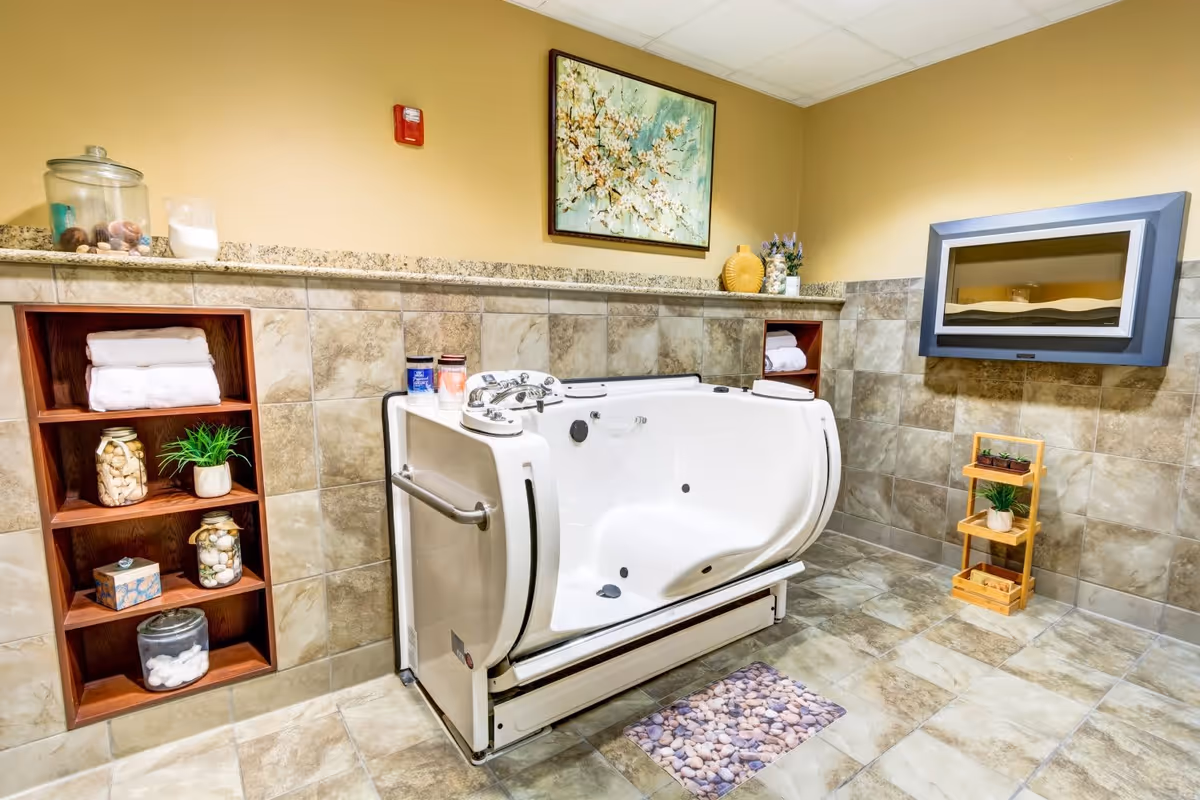 A spacious bathroom with a walk-in bathtub featuring safety handles and controls. The walls and floor are tiled in beige tones. There are built-in wooden shelves with towels, decorative jars, and plants. A framed floral painting hangs on the wall above the bathtub, and a modern electric fireplace is mounted on the adjacent wall. A small wooden plant stand with potted plants is placed near the fireplace.