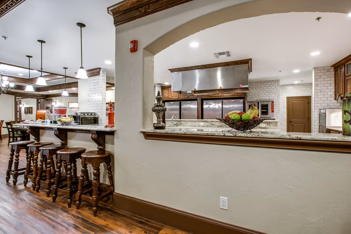 Interior view of a senior living facility kitchen and dining area with a long counter and wooden bar stools. The kitchen features stainless steel appliances, white tiled walls, and wooden cabinetry. The dining area has wooden chairs and tables, with pendant lights hanging from the ceiling.