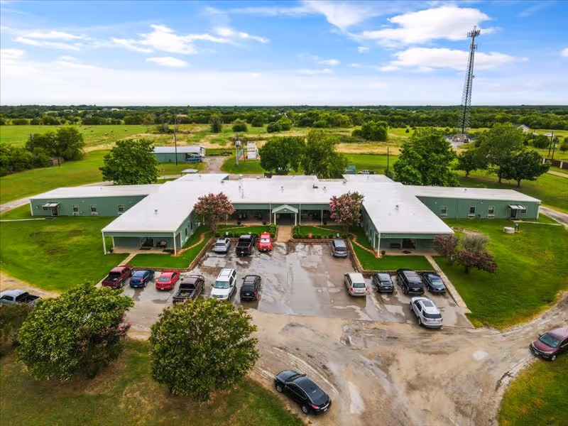 Aerial view of a single-story green building with a central parking lot, surrounding lawns and rural fields.
