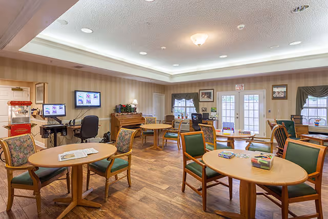 A well-lit common area in a senior living facility with multiple round wooden tables and green cushioned chairs arranged around the room. There is a piano against the far wall, a popcorn machine on the left, and two computer monitors on desks. The room has wood flooring, beige striped wallpaper, and windows with dark valances. French doors lead outside at the back of the room.
