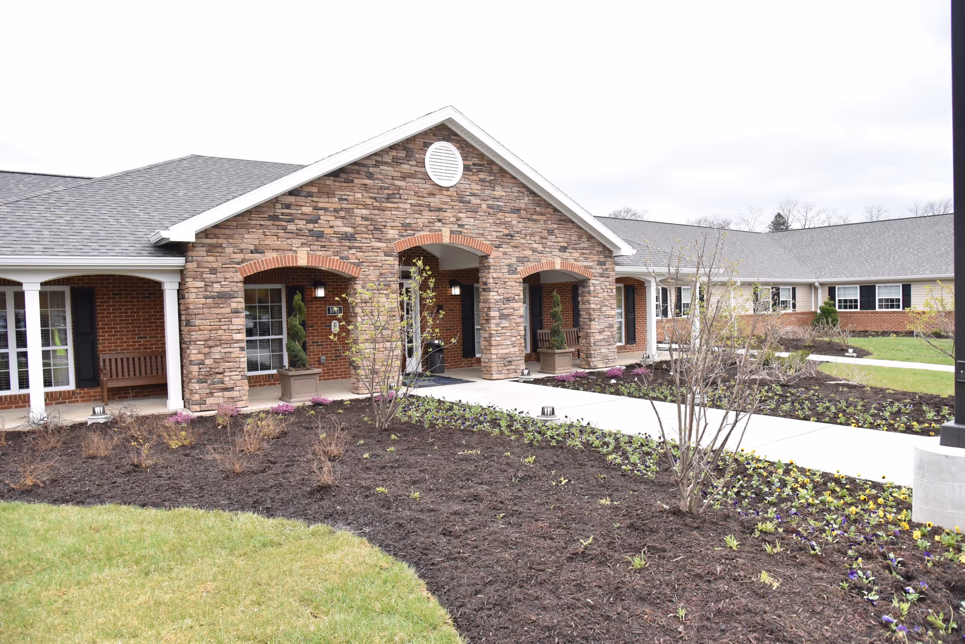 Exterior view of a senior living facility building with a stone and brick facade, a covered entrance with arches, potted plants, and landscaped garden beds with small plants and flowers along a concrete walkway.