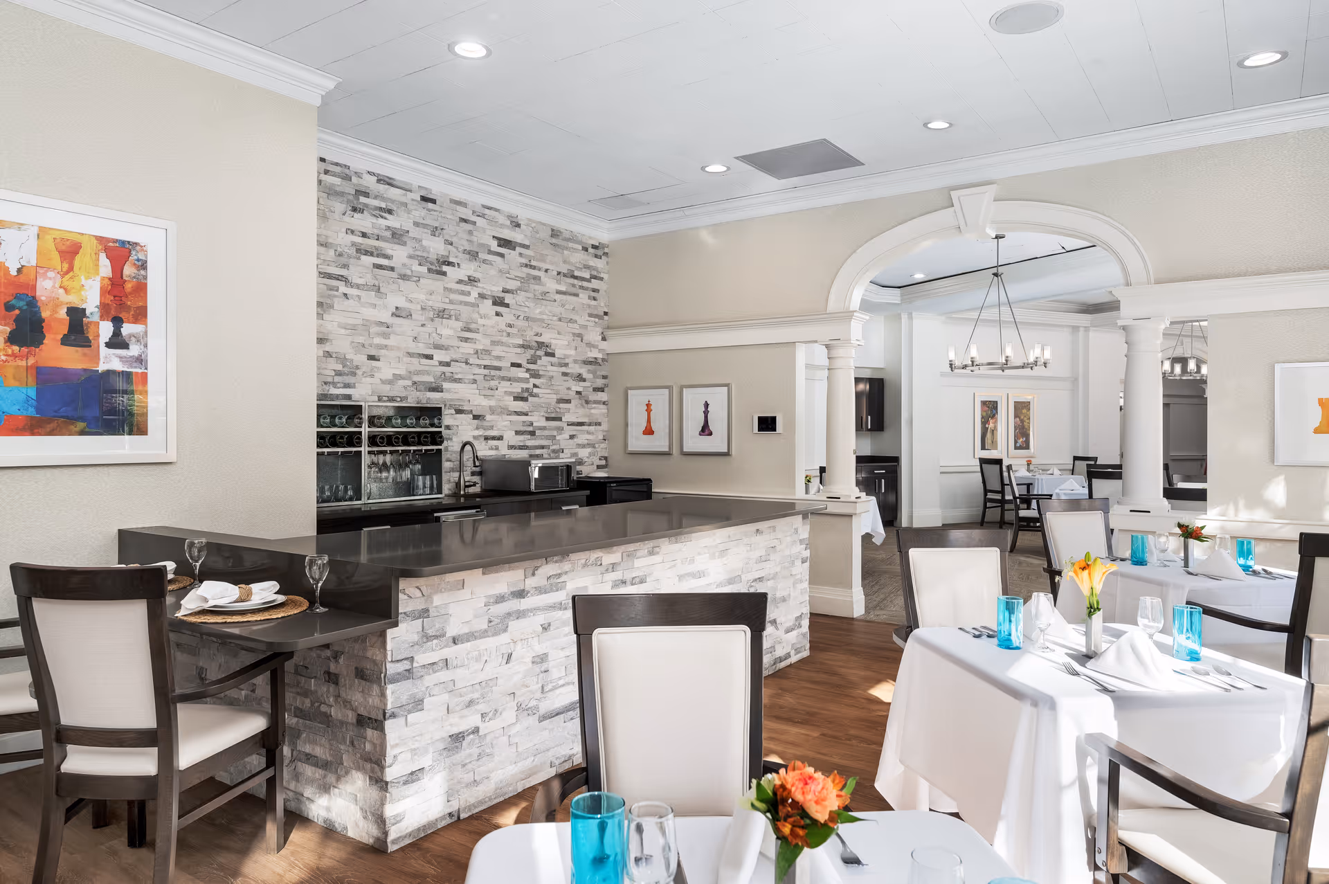 A bright dining area with tables covered in white tablecloths, set with blue glasses, white napkins, and floral centerpieces. There is a stone-tiled bar counter with a dark countertop and chairs around it. The room features light-colored walls, framed artwork, wooden flooring, and an arched doorway leading to another dining space with a chandelier.