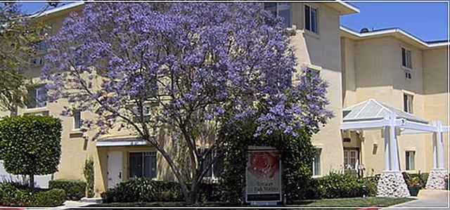 Exterior view of a multi-story senior living facility building with beige walls and several windows. In front of the building, there is a large tree with purple flowers and well-maintained shrubs and greenery. A sign near the entrance reads 'The Terraces at Park Marino'.