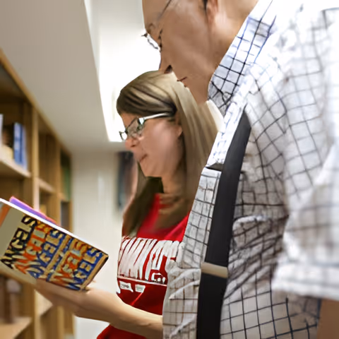 An elderly man and a younger woman standing side by side in a library or bookshelf area, both looking at a book the woman is holding titled 'Angels in America'.