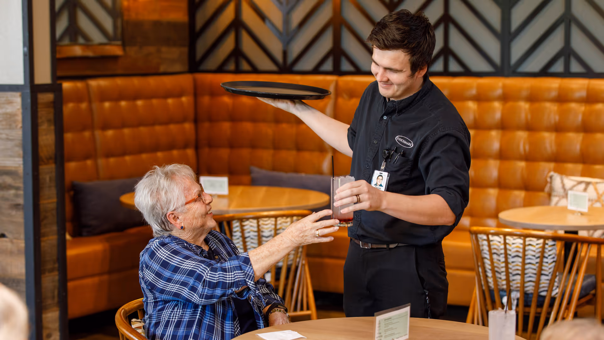 A young male server wearing a black shirt with a Touchmark logo hands a drink to an elderly woman seated at a table in a restaurant with brown leather booth seating and wooden chairs.