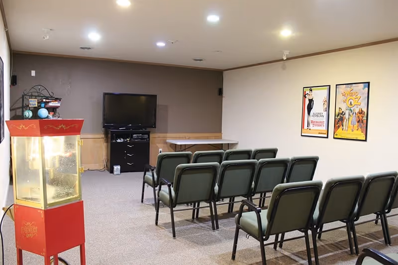 A small theater room with rows of green cushioned chairs facing a flat-screen TV mounted on a brown wall. There is a red popcorn machine on the left side and two framed movie posters on the right wall. The room has beige carpet and ceiling lights.