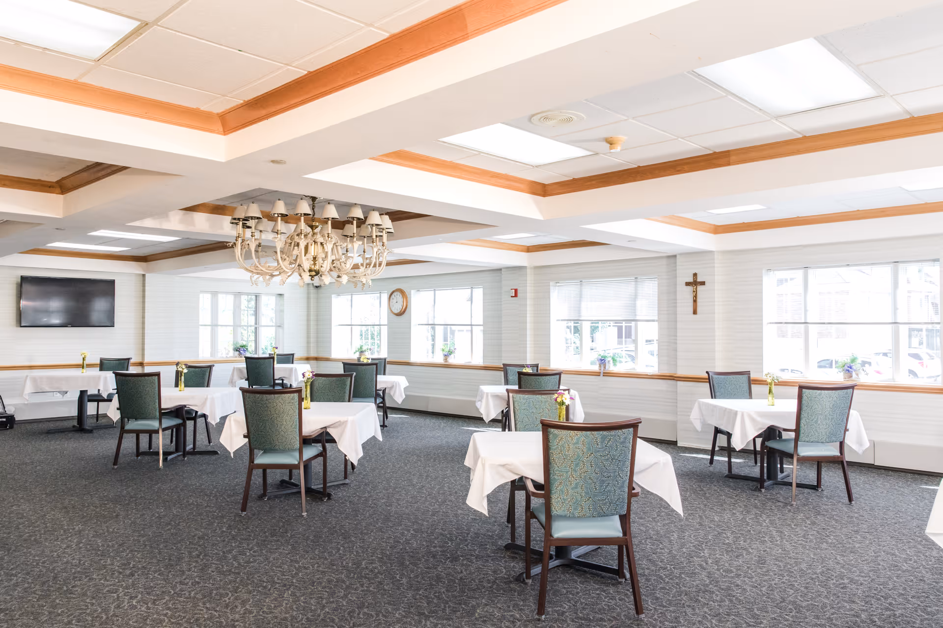 A bright dining room with several tables covered in white tablecloths, each with a small vase of flowers. The room has large windows letting in natural light, a chandelier hanging from the ceiling, a wall-mounted TV, and a clock on the wall. There is a cross mounted on one wall, and the carpet is dark with a subtle pattern.