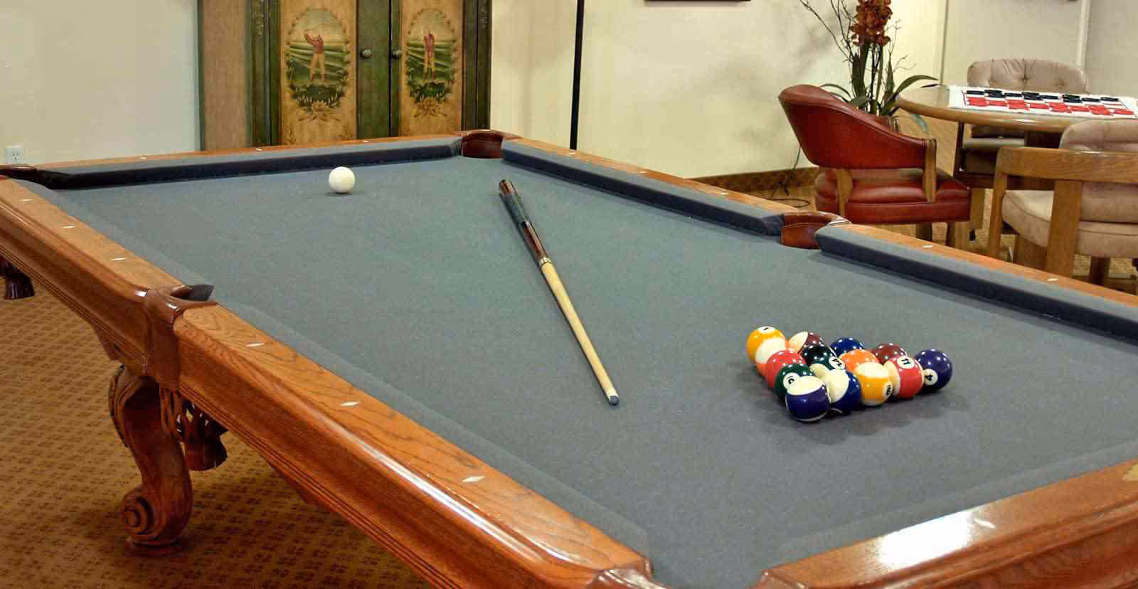 A billiards room with a pool table featuring a blue felt surface, a cue stick, a white cue ball, and a rack of colorful billiard balls. In the background, there are chairs and a table with a game set on it, along with a decorative folding screen with painted figures.
