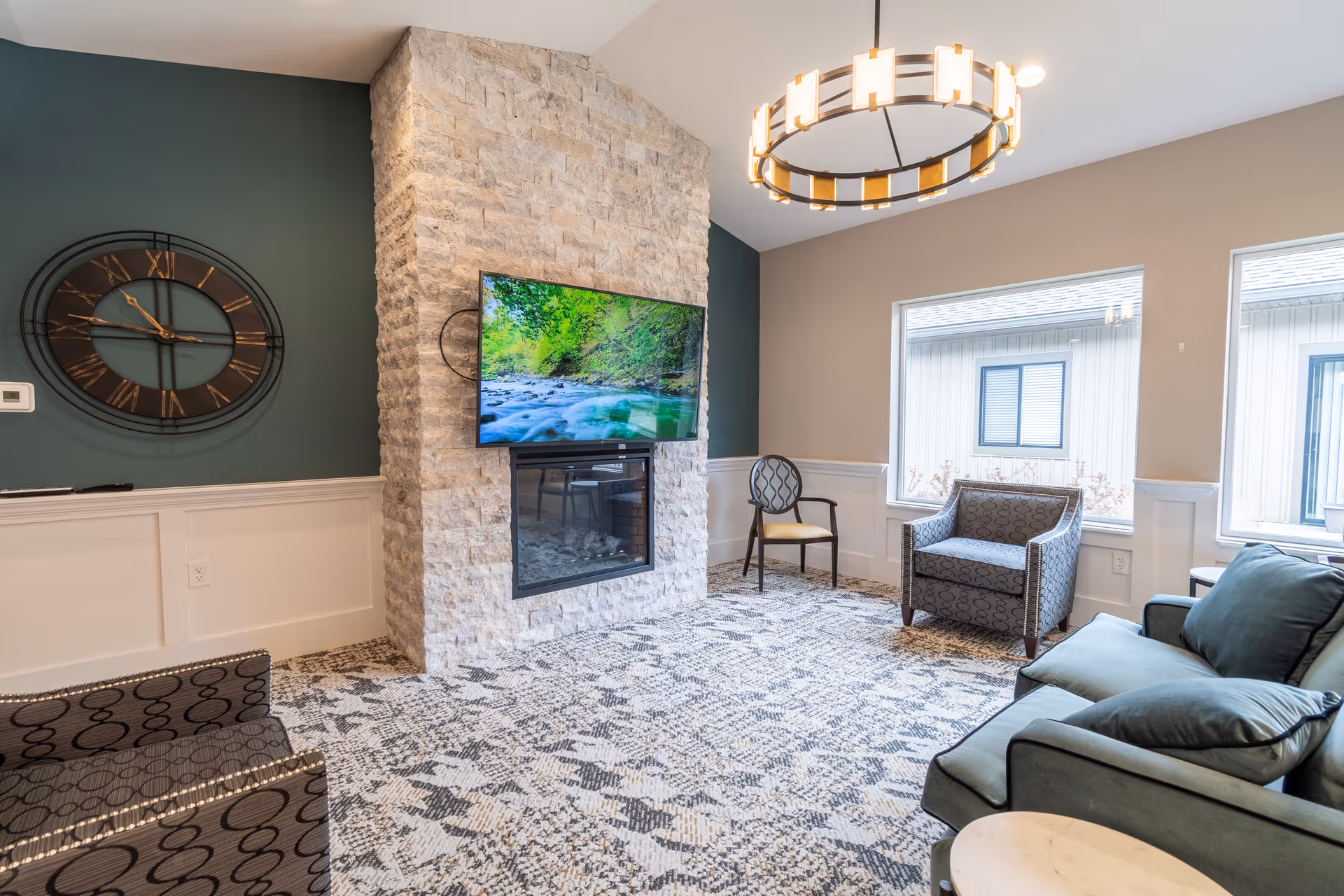 A cozy senior living facility common area with a stone fireplace, a wall-mounted flat-screen TV displaying a nature scene, a large round clock on a dark green accent wall, patterned carpet, and several chairs and a sofa arranged around the room. Large windows allow natural light to brighten the space.