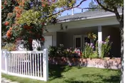 Front exterior view of a single-story building with a white picket fence, green lawn, flowering plants, and trees in front of a covered porch area.