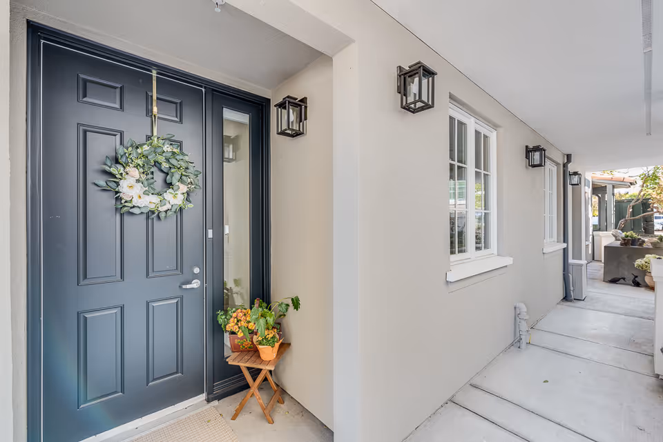 Entrance area of a senior living facility with a dark gray door decorated with a floral wreath. A small wooden table with potted plants is placed beside the door. The walkway is covered and lined with beige walls, windows, and black wall-mounted lantern lights.