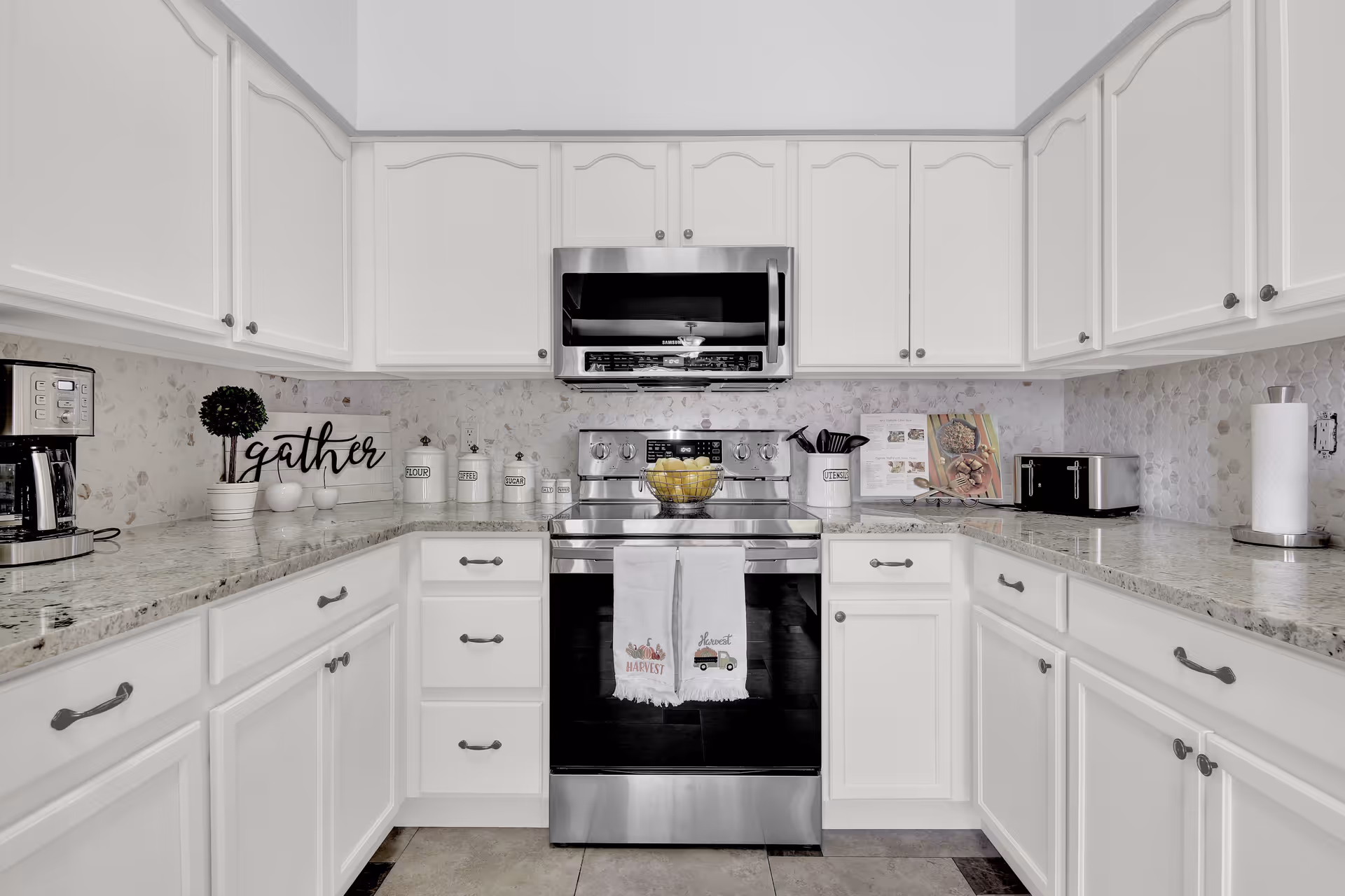 A modern kitchen with white cabinets and granite countertops. The kitchen features a stainless steel stove with a microwave above it, a coffee maker on the left counter, a toaster on the right counter, and various kitchen containers labeled flour, coffee, sugar, and utensils. Two decorative towels hang on the oven door, and a bowl of lemons sits on the stove. The backsplash has a light-colored hexagonal tile pattern.