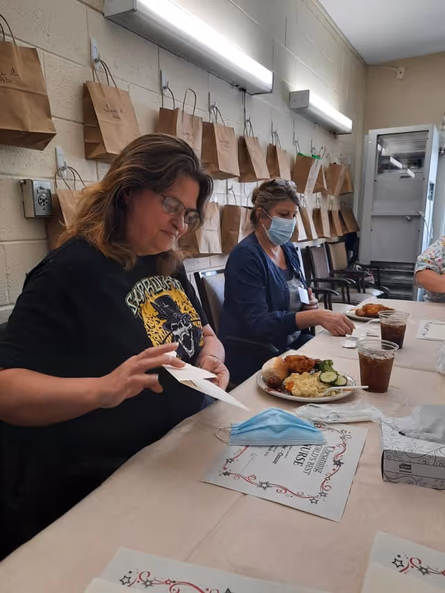Two women sitting at a table in a room with brown paper bags hanging on the wall behind them. One woman is wearing glasses and a black shirt with a graphic design, while the other woman is wearing a face mask and a blue jacket. On the table are plates with food, drinks, a face mask, and a tissue box.