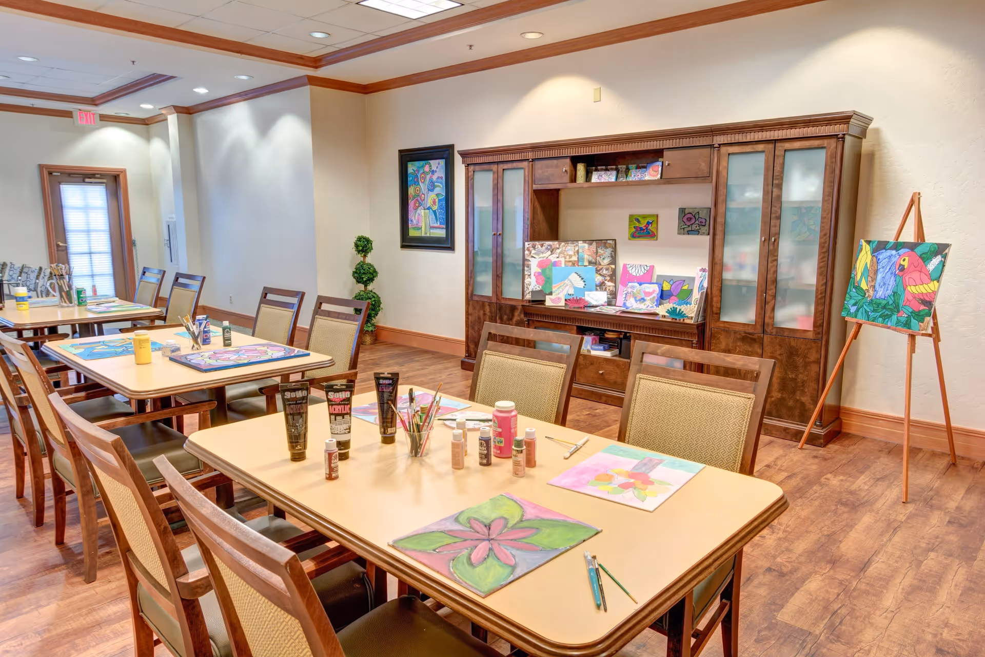 Well-lit community activity/dining room with tables and chairs set up for painting, art supplies on the tables, and finished artwork displayed on a cabinet and easel.