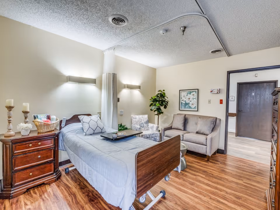 A well-lit senior living facility room with a hospital-style bed covered in gray bedding, a wooden nightstand with candles and a basket, a floral armchair, a small gray couch, a potted plant, and a framed floral painting on the wall. The room has wood flooring and a doorway leading to another area.