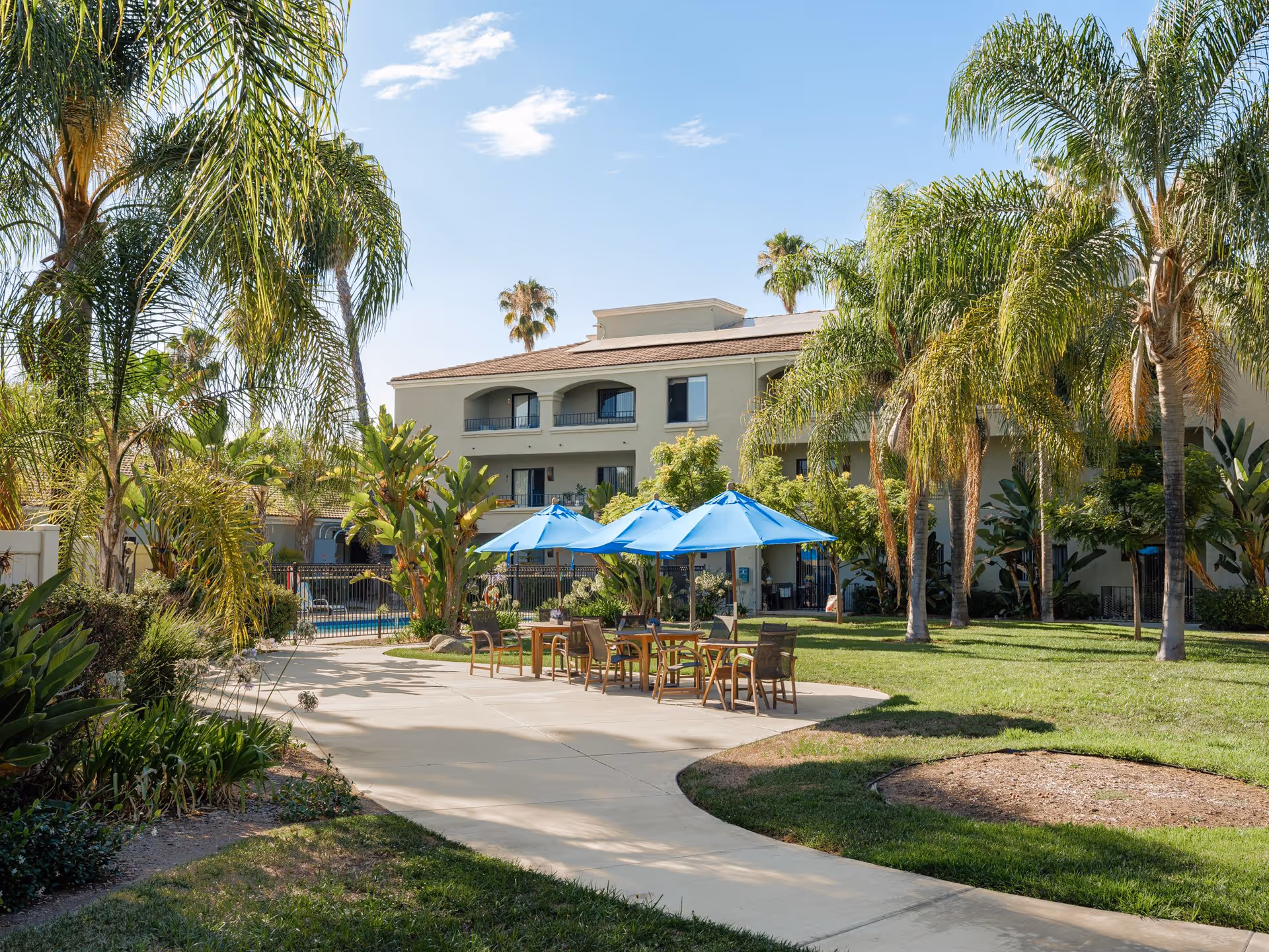 Outdoor area of Las Villas Del Norte featuring a paved walkway, green lawn, palm trees, and several tables with blue umbrellas. In the background, there is a two-story building with balconies and windows under a clear blue sky.